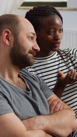Interracial couple talking and waving on video call conference using modern smartphone, sitting on couch. Multi ethnic people with technology chatting using online remote communicationの写真素材