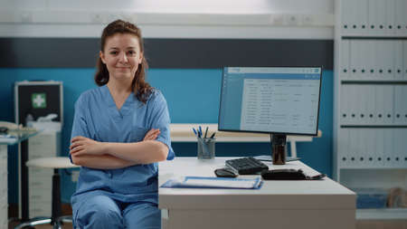 Portrait of woman working as medical assistant at desk in doctors office. Nurse looking at camera and smiling while preparing for work on computer. Healthcare specialist in uniformの写真素材
