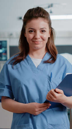 Portrait of medical assistant holding checkup documents to help doctor with treatment and medicine. Woman working as nurse with uniform and tools looking at camera to cure patientの写真素材