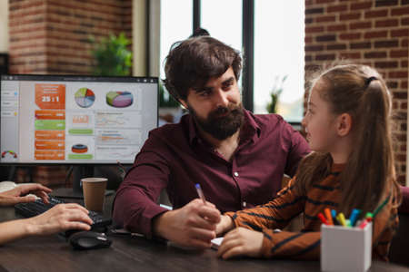 Company office young worker talking with little girl while sitting at desk and smiling. Child conversating project manager business man while in modern agency sitting at table.の写真素材