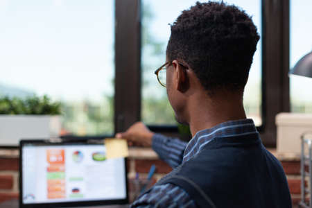 Over shoulder view of startup owner working at desk sticking yellow post it on corner of laptop...