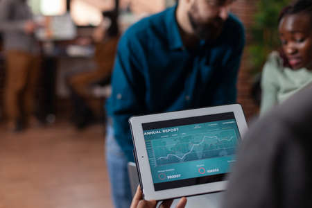 Multiethnic businesspeople holding tablet computer with company turnover on screen working at marketing project discussing business strategy working at startup office. Diverse team brainstorming ideasの写真素材