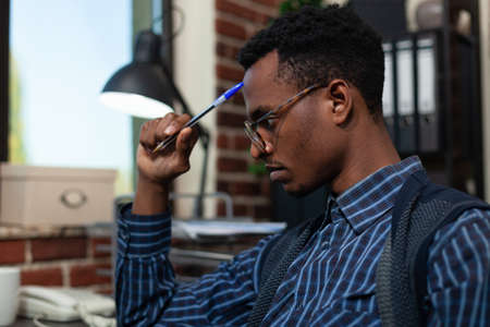 Portrait of startup employee touching forehead with pen looking focused at laptop screen with...