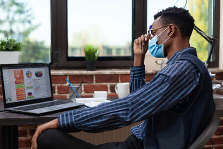 Uninspired entrepreneur wearing face mask feeling tired looking at laptop screen with business...