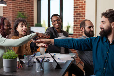 Happy successful multiethnic businesspeople putting hand on hand enjoying business achievements during marketing meeting in startup office. Diverse team working at company turnoverの写真素材