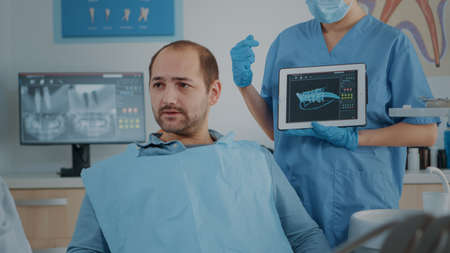 Dentistry assistant showing x ray scan results on tablet to patient, explaining cavity problems and...