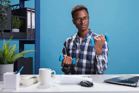 Fit and athletic man doing fitness training exercise for stiff muscles. Active confident and strong young adult lifting dumbbells in order to stay in shape while doing office work.の写真素材