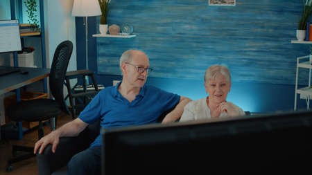 Elder couple watching movie on tv to enjoy retirement in living room, having fun with comedy on television. Senior people sitting on sofa and looking at comedy channel on screen to have fun.の写真素材