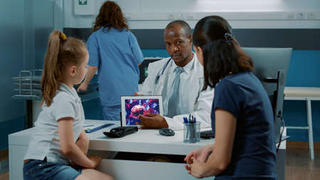 Doctor explaining coronavirus illustration on tablet display to adult and little child in medical...
