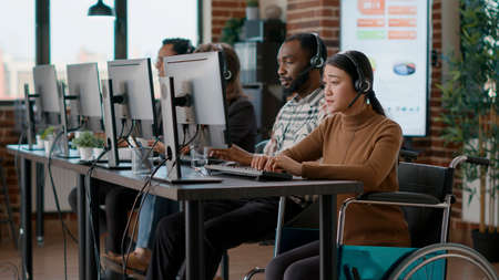 Asian woman with disability using headset to help clients at call center, talking to people about...
