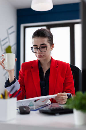 Businesswoman with glasses holding cup and looking at clipboard with chart. Startup owner in red jacket reading business papers while having a coffee. Employee working realxed drinking tea.の写真素材