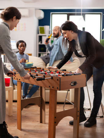 Multiethnic group of people playing at foosball game table, enjoying soccer match with toy in...