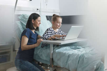 Joyful mother enjoying leisure time with smiling cheerful daughter while in pediatric clinic ward room using laptop. Mother and daughter sitting in pediatric hospital room while using computer.の写真素材