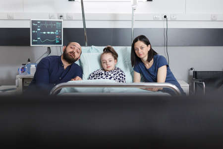Parents and ill little daughter watching television family program while sitting in pediatric hospital ward treatment room. Family spending time together in clinic patient room while enjoying tv show.の写真素材