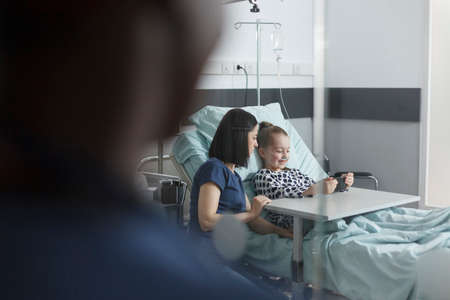 Cute little girl playing on smartphone while caring young mother sitting beside patient bed in pediatric clinic room. Joyful patient enjoying mobile phone entertainment while sitting in recovery ward.の写真素材