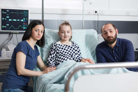 Portrait of sick little girl laying in hospital bed with worried uneasy parents beside her while looking at camera. Caucasian family in pediatric clinic patient recovery ward room.の写真素材