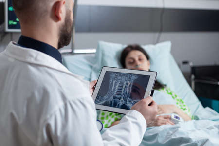 Medic holding digital tablet with throat mri looking at sleeping patient in hospital bed after throat surgery. Woman resting connected to screen measuring vitals after surgical intervention.の写真素材