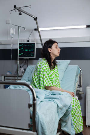 Bored woman recieving iv drip line treatment sitting on bedside looking into distance in hospital ward. Patient with illness feeling depressed waiting for doctor to do clinical consultation.の写真素材