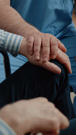 Close up of hands of nurse and senior patient at medical visit in nursing home. Medical assistant giving support and comforting old man in wheelchair. Specialist helping disabled personの写真素材