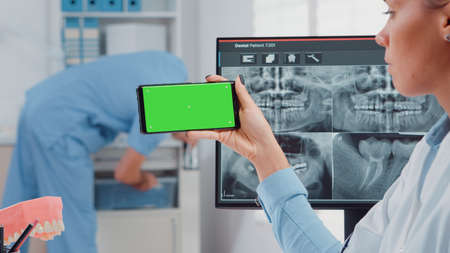 Close up of dentist analyzing smartphone with green screen and teeth scan on computer in dental cabinet. Woman holding device with chroma key for isolated mockup template for dentistryの写真素材