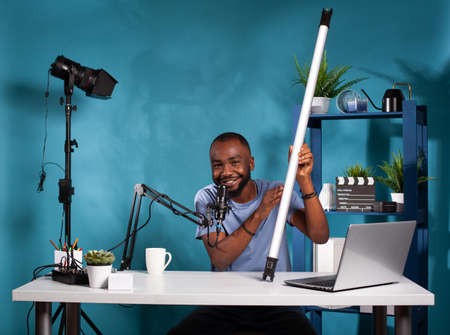Vlog reviewer presenting rgb tube video light sitting at desk with laptop in vlogging studio recording podcast. Vlogger testing videography continous lighting equipment for social media channel.の写真素材