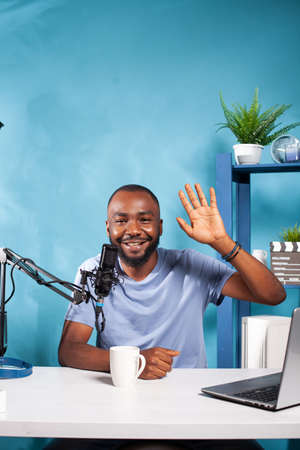 Portrait of african american vlogger waving hand at camera in recording studio with professional...