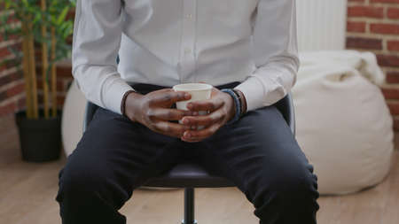 Close up of man with cup of coffee in hands attending aa therapy meeting. Nervous adult with alcohol addiction having beverage and talking to people at rehabilitation program session.の写真素材