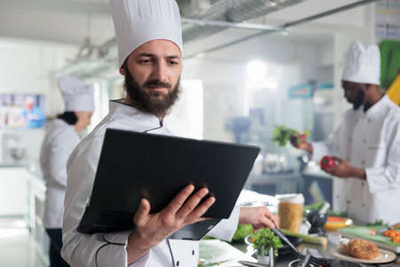 Food industry worker having laptop, following gourmet dish recipe on screen stirring ingredients in pan. Head chef with computer preparing garnish for main course while in restaurant kitchen.の写真素材