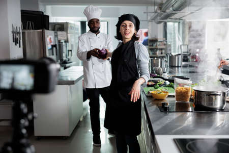 Head chef recording cooking session while getting help from sous chef while cooking gourmet dish. Cuisine cooks in restaurant professional kitchen filming culinary course for internet broadcast.の写真素材