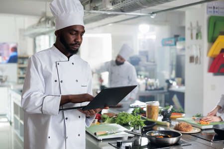 Gourmet cuisine head chef with laptop looking for gastronomic dish recipes and garnish ideas in restaurant kitchen. Gastronomy expert wearing professional cooking uniform while searching meal ideas.の写真素材