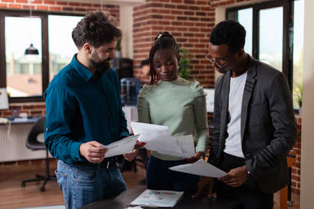 Multiracial team holding papers analyzing marketing charts discussing business project in brick wall office. Multi-ethnic businesspeople brainstorming ideas working at company strategy. Corporate staffの写真素材