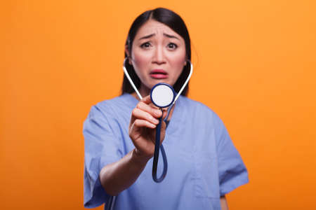 Agitated uneasy asian nurse using stethoscope to consult sick patient on orange background. Distressed caretaker with professional medical instrument consulting sick person.の写真素材