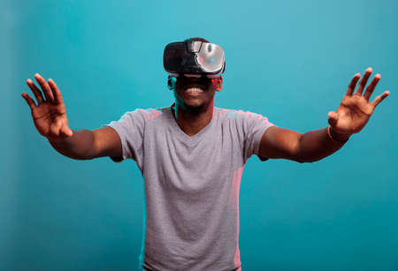 Cheerful guy wearing vr goggles with video interaction, standing over blue background. Young person using virtual reality technology with 3d tech simluation and futuristic visual.の写真素材