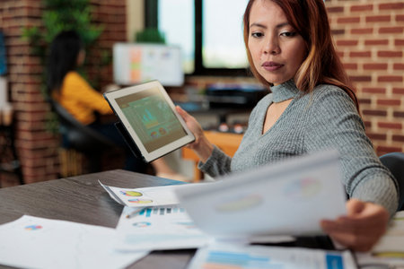 Asian manager holding tablet computer checking management statistics working at company strategy in startup office. Businesswoman analyzing papers with graph planning partnership projectの写真素材