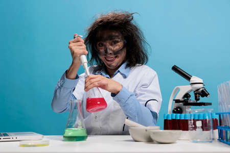 Crazy looking mad chemist grinning creepy at camera while using dropper to to laboratory work after chemical explosion. Foolish silly female lab worker with pipette sitting at desk on blue background.の写真素材