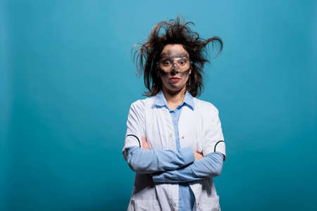 Humorous mad scientist with erratic hair and filthy face standing with crossed arms. Hilarious looking crazy chemist with messy hairstyle and dirty face standing on blue background looking at camera.の写真素材