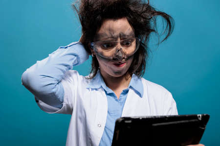 Wacky and silly looking crazy chemist with touchscreen tablet, puzzled by failed experiment report. Mad scientist with messy hair and dirty face looking confused after dangerous chemical explosion.の写真素材