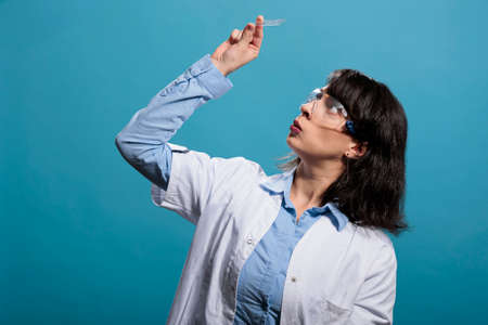 Young adult curious microbiology researcher analyzing bacteria experiment results on microscope glass slide. Biochemistry specialist looking at glass tray while on blue background. Studio shot.の写真素材