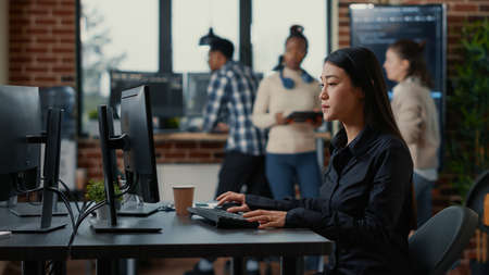 Asian software developer holding clipboard sitting down and starting to type on computer keyboard. Mixed team of programmers working together on analyzing code on wall screen tv.の写真素材