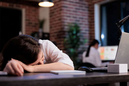 Fatigued marketing agency employee sleeping on desk because of overtime working hours at night. Exhausted company employee falling asleep in office workspace while working on project.の写真素材
