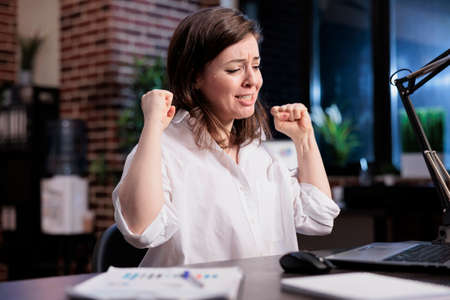 Enthusiastic positive young adult entrepreneur being excited about new job while in office at night. Happy joyful young adult businesswoman celebrating finishing startup project before deadline.の写真素材