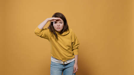 Curious woman looking into the distance far away searching for something with palm over forhead in studio. Young person scouting the horizon seeking for someone over yellow background.の写真素材
