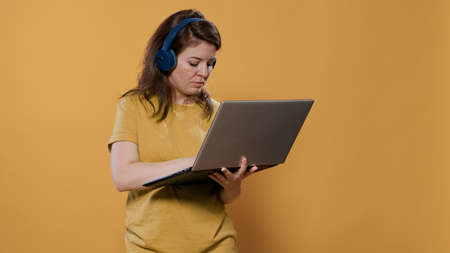Casual woman wearing wireless headphones holding laptop and typing on keyboard looking at screen standing in studio. Relaxed person listening to music on earphones using portable computer.の写真素材