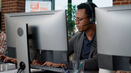 African american employee using headset at customer service job, asnwering call about telemarketing sales. Male operator working at call center office to help clients on helpline.の写真素材
