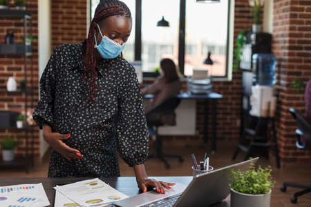 Pregnant businesswoman wearing virus protection mask in office using work laptop to review marketing strategy and proposed project ideas. Office worker reviewing accounting documentation.の写真素材