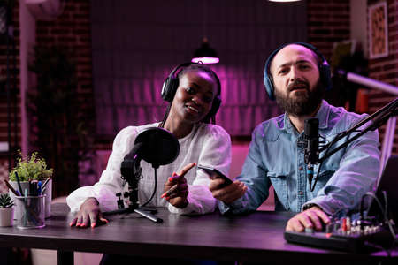 Male presenter and guest recording podcast vlog discussion in studio with neon lights. Multiethnic...