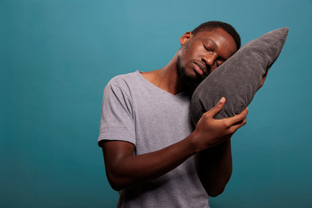 Tired man using comfortable pillow to get sleepy and take nap over blue background. Exhausted person...