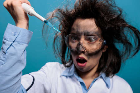 Crazy mad chemistry reasearcher with dirty face and messy hair experimenting with unknown compounds. Lunatic chemist looking shocked at professional pipette filled with experimental liquid substance.の写真素材