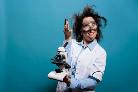 Mad foolish scientist with messy hair and dirty face having professional microscope while acting goofy on blue background. Silly looking chemist looking dumb after laboratory experiment explosion.の写真素材