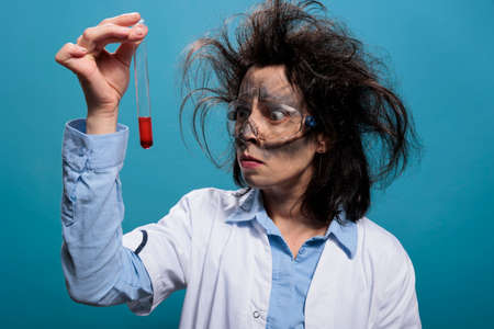 Mad worried chemist with messy hair and dirty face frowning at flask filled with red liquid substance sample. Insane anxious looking scientist analyzing chemistry glass tube filled with blood sample.の写真素材